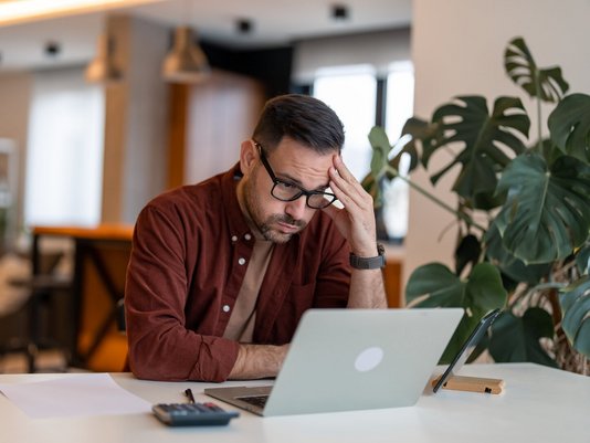 Verzweifelter Bauherr am Tisch mit Laptop, Taschenrechner und Papieren – steht sinnbildlich für finanzielle Belastung durch Bau-Insolvenz ohne passenden Schutz beim Hausbau