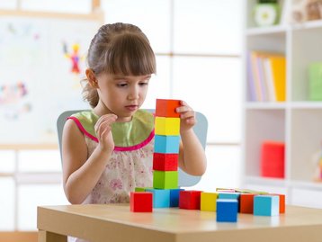 Mädchen stapelt erste Bausteine konzentriert, symbolisiert gesicherte Bauphase mit Qualität, 20 Prozent Fertigstellungsbürgschaft und Treuhand Baugeldkonto